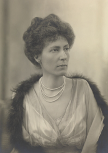 Sepia image of a woman wearing a light coloured dress, three pearl necklaces, and a thin dark feather or fur bower draped across her shoulders. She is looking to her left without expression.