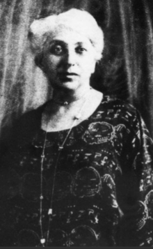 A middle-aged white woman with white hair and dark eyes, wearing a dark print dress with a round neckline