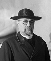 A bespectacled Portuguese man with facial hair wearing a black galero without tassels and black overcoat.