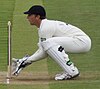 A young man with dark hair is dressed in an all white cricket kit with a blue peaked cap, large gloves and thick pads covering his lower legs. He is crouched low behind a set of wooden stumps.