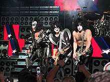 The band Kiss is shown onstage at a concert. From left to right are the bassist Gene Simmons, two electric guitarists and the drummer, who is at the rear of the stage. Simmons is wearing spiked clothing and his tongue is extended. All members have white and black face makeup. Large guitar speaker stacks are shown behind the band.