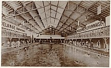 Vue du grand bassin de la piscine avec des nageurs sous un toit du type halle. Au fond du bassin sur le petit côté se trouvent des gradins.