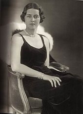 Photographie en noir et blanc d'une jeune femme assise, portant des cheveux courts et une tenue de soirée noire.