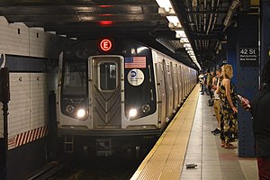 An E train, here composed of R160A cars is seen entering the 42nd St-Port Authority Bus Terminal station. The front of the train contains two white lights providing slight illumination, a window on the left side, the American flag on the right side, and the MTA logo below the flag.