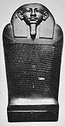 Black and white image of a dark stone coffin with a human face, the coffin stands upright facing the viewer.