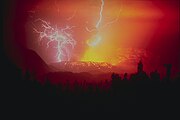 A photograph depicting lightning striking a volcano that is in the process of erupting bright yellow lava into the air, all surrounded by a red haze.