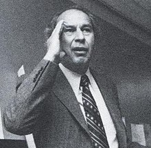 Lois holding his hand to his head, wearing a suit in a black-and-white photograph