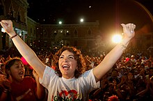 La imagen muestra a Ivonne Ortega levantando ambos brazos en señal de celebración. Porta una playera blanca que con letras rojas dice "Ganamos".