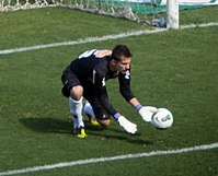 Homem de pele clara e cabelo preto curto, vestindo um uniforme de futebol com camisa e calções pretos e meias brancas, agachado em frente a um gol com suas mãos enluvadas esticadas em direção a uma bola de futebol em movimento.