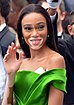 The image features a woman (Winnie Harlow Cannes) with a smiling expression, waving her hand. She has vitiligo, characterized by patches of skin with depigmentation. Her hair is styled straight and parted to the side. She is wearing makeup with prominent eyelashes and eye shadow. She is dressed in an off-the-shoulder, vibrant green garment. Earrings are visible, and the setting is an event with other people in the background.