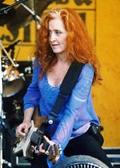 A woman with curly red hair wearing a blue shirt holding a guitar.