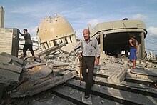 A man with a walking stick walks amongst the ruins of a mosque. In the background a child stands looking at the ruins, while a man traverses the rubble.