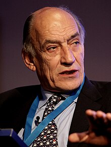 The head and shoulders of a white man in a dark suit and tie with some grey hair, predominately bald