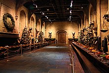 Une très grande salle de banquets au sol de pierre et haute de plafond, dans laquelle sont disposées deux longues tables contre les murs latéraux, et des sapins de noël à intervalles réguliers