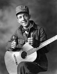 Black and white promotional portrait of a man (Jimmie Rodgers) dressed in a 1930s railway man attire consisting of a denim jacket, denim pants, a cap, and a bowtie with a white shirt. He gives thumbs up while he holds a guitar with his forearms on his raised left knee and standing on his right leg