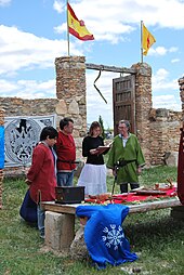 Four individuals stand by an outdoor table. A tall stone wall is visible in the background.