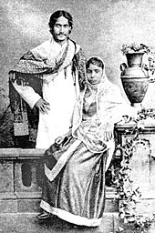 Black-and-white photograph of a finely dressed man and woman: the man, smiling, stands with the hand on the hip and elbow turned outward with a shawl draped over his shoulders and in Bengali formal wear. In front of him, the woman, seated, is in an elaborate dress and shawl; she leans against a carved table supporting a vase and flowing leaves.