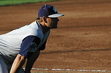 A man wearing a sleeveless gray baseball jersey with blue undershirt and a blue cap crouching