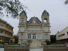 Photographie d'une villa en pierre taillée claire, à plusieurs étages et haut toit d'ardoises, flanquée de deux clochetons de chaque côté. Accès par un escalier monumental à double révolution en pierre et hautes fenêtres aux huisseries blanches avec des balcons à colonnades.