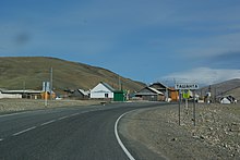 Route avec le panneau d'arrivée à Tachanta. En second-plan, les maisons du village, le tout dans un paysage aride.