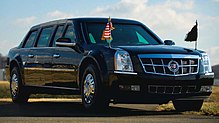 A black Cadillac limousine is at a 45-degree angle, showing to the camera its front and starboard sides. Two small flagpoles are mounted to the front fenders, one flying a US flag with gold fringe.