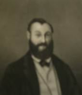 A sepia-toned, historical portrait of a bearded man wearing a suit and bow tie, with an indistinct facial expression.