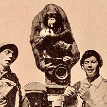 Orangutan perched on top of 1916 film camera, young men on either side