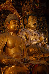 Three-quarter view of two cross-legged seated statues (one in front of the other). The statue in the back appears larger and shows the Vajra Mudrā, enclosing the index finger of the left hand with the fist of the right hand. The statue in front has both hands resting on the lap. Color photograph.