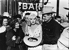 Monroe holding a hat and standing in the middle of a crowd of people, facing the camera. On her right is Gable and on her left, Winwood. There is a sign that says 'BAR' in the background.