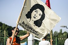 A woman demonstrator in sunglasses waves a white flag depicting the face of murdered trade unionist Marsinah. Indonesian language text appears below Marsinah's face