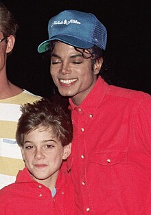 A smiling Jackson wears a blue baseball cap and a red shirt. On his left, a young boy smiles. He is dressed in a red shirt, too.