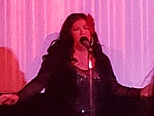 Photographie d'une femme qui chante devant un micro, habillée en noir.