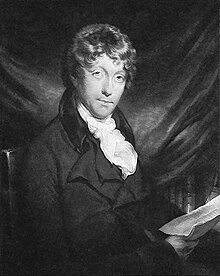 A young man with brown, curly hair sitting in front of some kind of cloth. Books can be seen underneath the cloth; the man is wearing a jacket with a white cravat