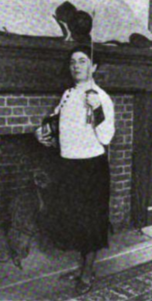 A white woman standing before a brick hearth, holding a fencing foil