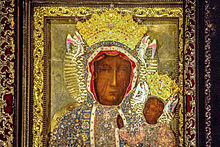 Portrait of a black skinned woman, wearing a black printed cloth around her body and head. She is holding up a baby in her left hand. Both of them have a yellow halo behind them.