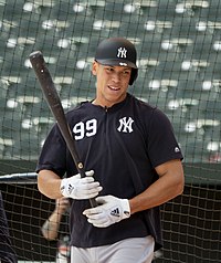 Medium-wide shot of Aaron Judge holding a bat and wearing a "NY" shirt and batting helmet.