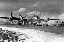 Black and white photo of a four engined propeller aircraft sitting on tarmac. Scaffolding has been erected near the nose of the aircraft and men are working on equipment nearby. The tail of another aircraft is visible to the right of the photo and buildings are visible behind the main aircraft.