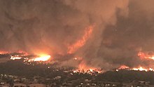 A vortex of brown smoke rises and mixes with flames in a helicopter view of a suburb