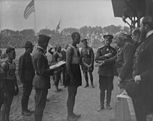 A black-and-white photograph of a man surrounded by military people and receiving a medal from an older military man