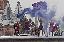 Eight women wearing colorful summer clothes and knit ski masks stand on a snowy stage. One is playing a guitar, another waves a purple flag with a combination woman symbol-raised fist, and another holds a smoke bomb emitting a purple-blue plume of smoke.
