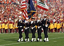 Photograph of five members of the Naval Base Ventura County Honor Guard parading the colors, the University of Southern California Marching Band is in the background wearing yellow and performing the national anthem during the 2008 Rose Bowl game