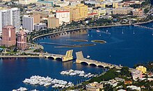 A bridge connects a dense urban area with a slightly less dense one. Several rows of boats are moored near the latter. Both sides of the bridge are supported by arches. The middle segments of the bridge are raised almost fully vertical. A boat has just passed through it, heading away from the moored boats.