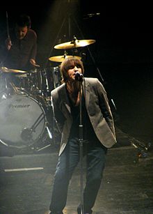 A brown-haired man sings into a microphone; in the background, another man plays drums.