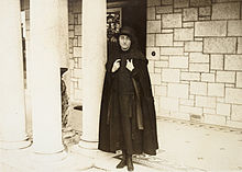 Woman in a black cape, drop-waist dress and a hat, standing beside three pillars in front of a block wall