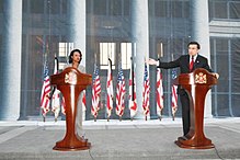 Woman and man standing in front of podiums, with flags in background