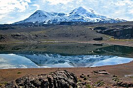 A snow-covered mountain with two hump-like summits rising above an unvegetated landscape with a lake