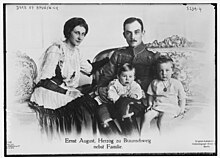 Carte postale en noir et blanc montrant un couple assis sur des fauteuils avec deux garçonnets ; le père porte un uniforme allemand, la mère une robe avec des broderies.