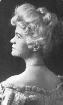 A middle-aged white woman with blonde or white hair in a bouffant updo, photographed in profile, wearing a frilly off-the-shoulder dress