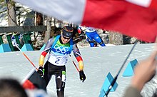 A woman in multicoloured winter sportswear which features the Olympic rings on her cheast and the number 2 in the center, moves twoards the camera. She is pictured in an area covered in snow. A second person can be seen in the background to her right. An out of focus red-and-white flag covers the upper parts of the image.