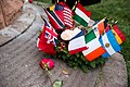 Photo agrandie de la base grise du monument. Une couronne de houx verte et entourée de plusieurs drapeaux de divers pays et de fleurs.